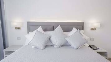 Una cama bien hecha, con cabecera gris, almohadas blancas y una mesita de noche con lámpara y teléfono.