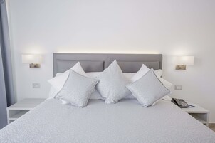 Una cama bien hecha, con cabecera gris, almohadas blancas y una mesita de noche con lámpara y teléfono.