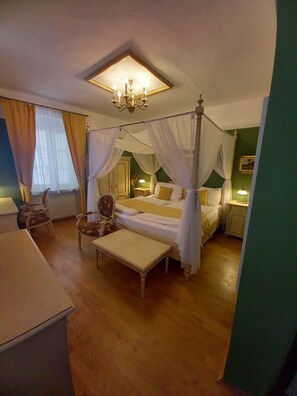 Una cama con dosel sobre cuatro columnas, con una mesita de noche, una silla y una mesa en una habitación con suelo de madera y paredes verdes.