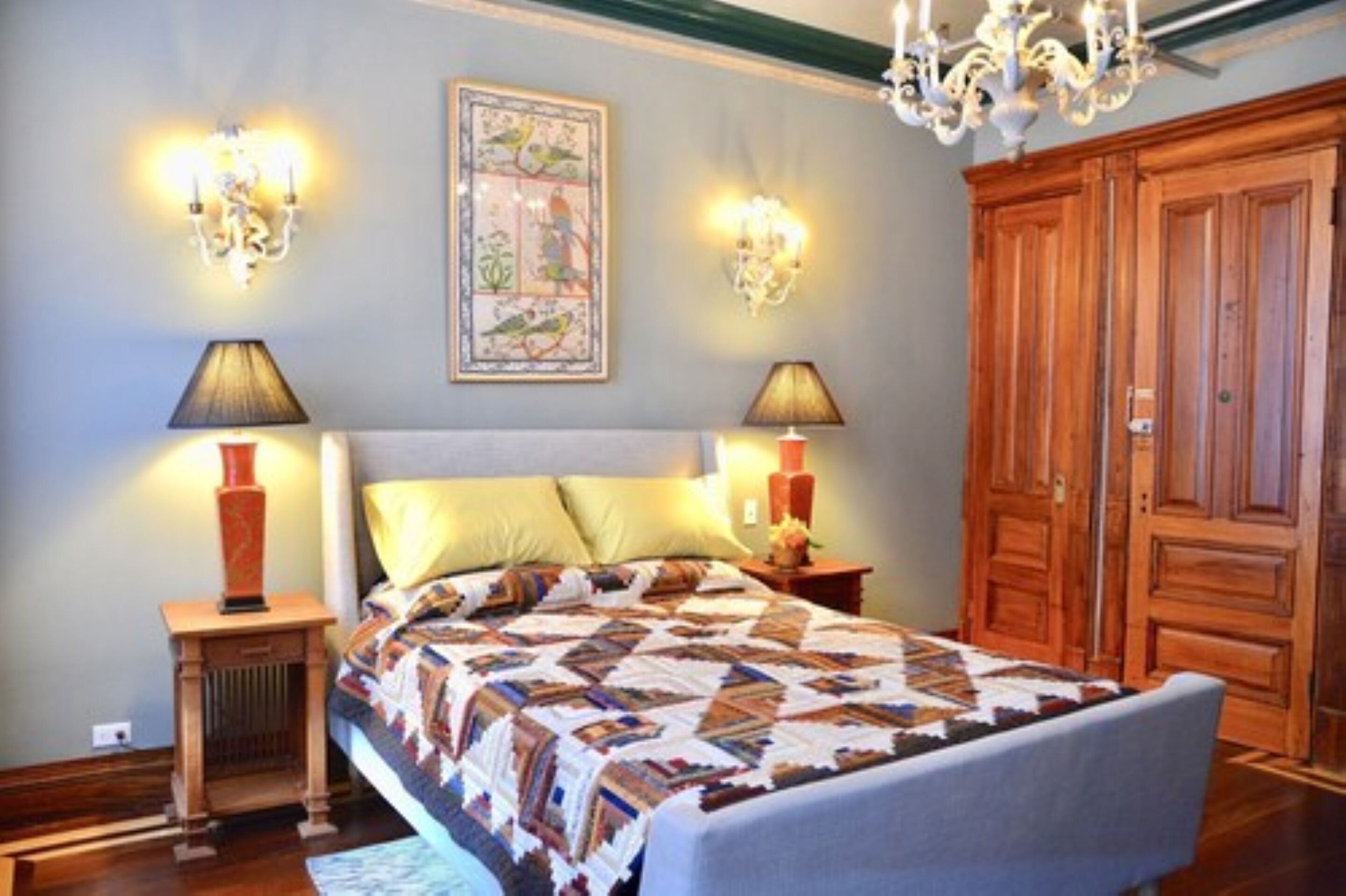A bedroom with a large bed, two wall-mounted lamps, a chandelier, and a wooden wardrobe.
