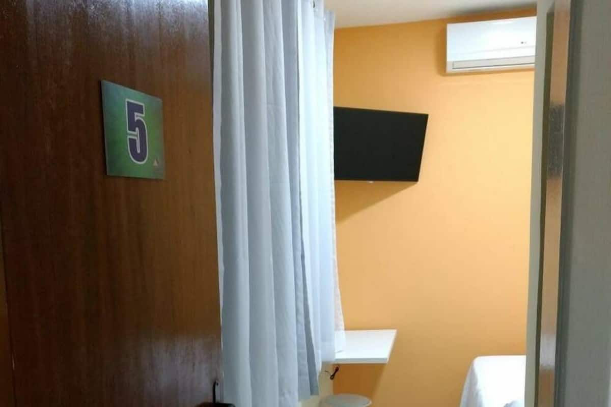 Une petite pièce, de taille modeste, avec une porte en bois portant le numéro 5, un rideau blanc, une petite table blanche, un lit recouvert d’un drap blanc et un appareil de climatisation fixé au mur.