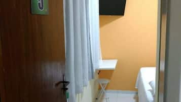 A small, compact room with a wooden door marked with the number 5, a white curtain, a small white table, a bed with a white sheet, and an air conditioning unit on the wall.