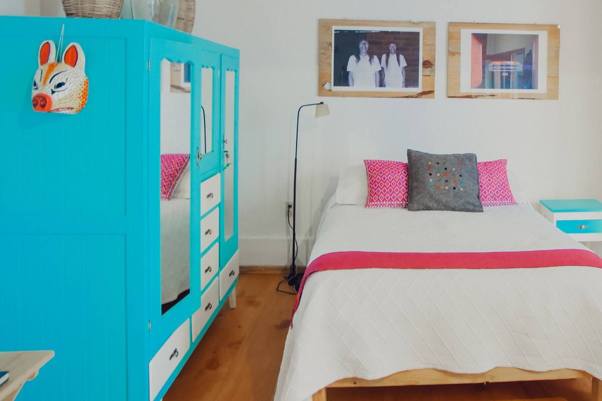 Een slaapkamer met een turquoise garderobe, een bed met een witte quilt, roze en grijze kussens, en twee ingelijste foto's aan de muur.