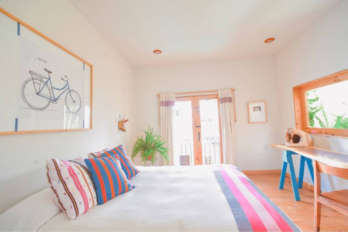 Een slaapkamer met een bed, kleurrijke kussens, een bureau, een tekening van een fiets op de muur en een raam met uitzicht.
