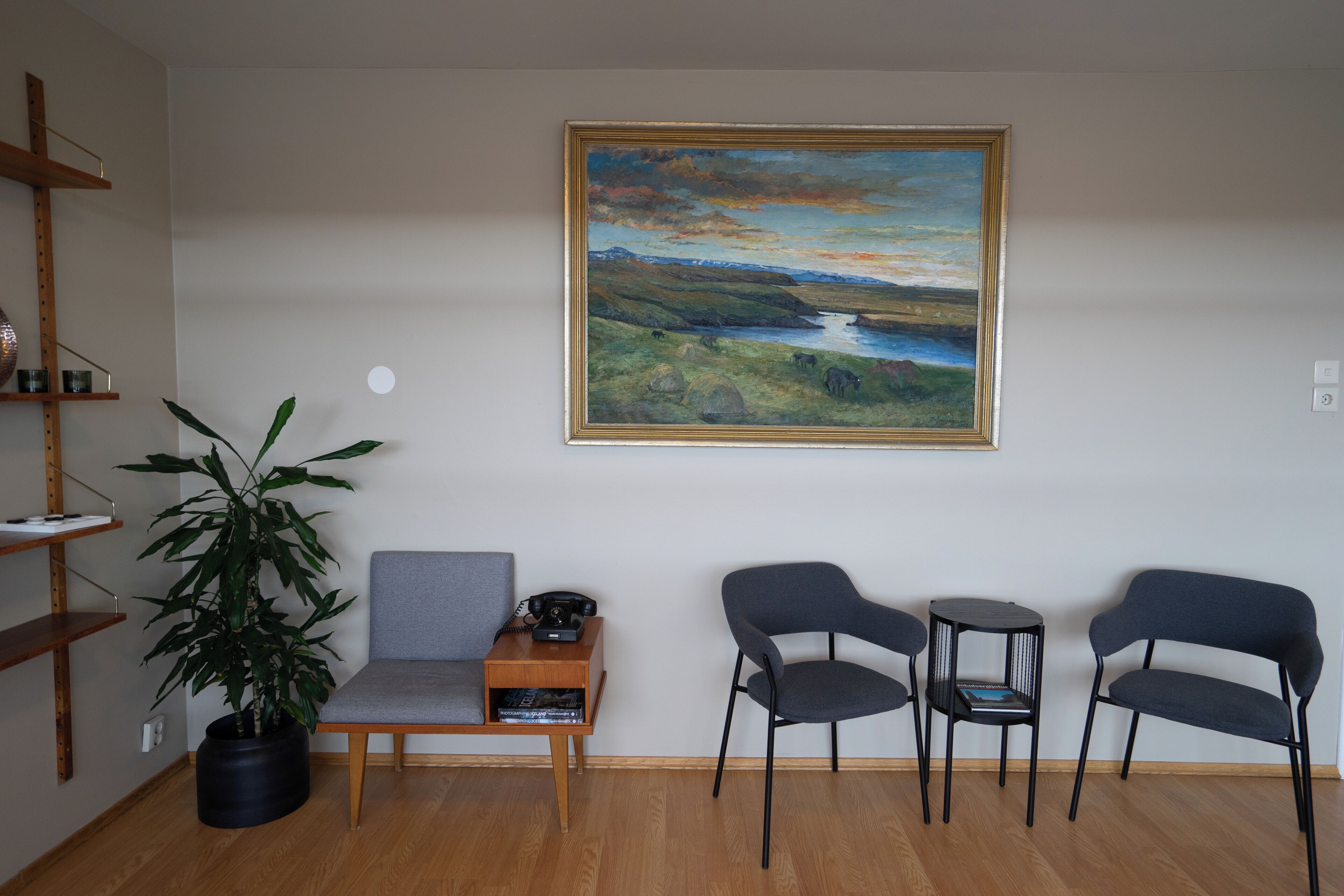 A room with a painting of a landscape, a sofa, two chairs, a small table, a potted plant, and a bookshelf.