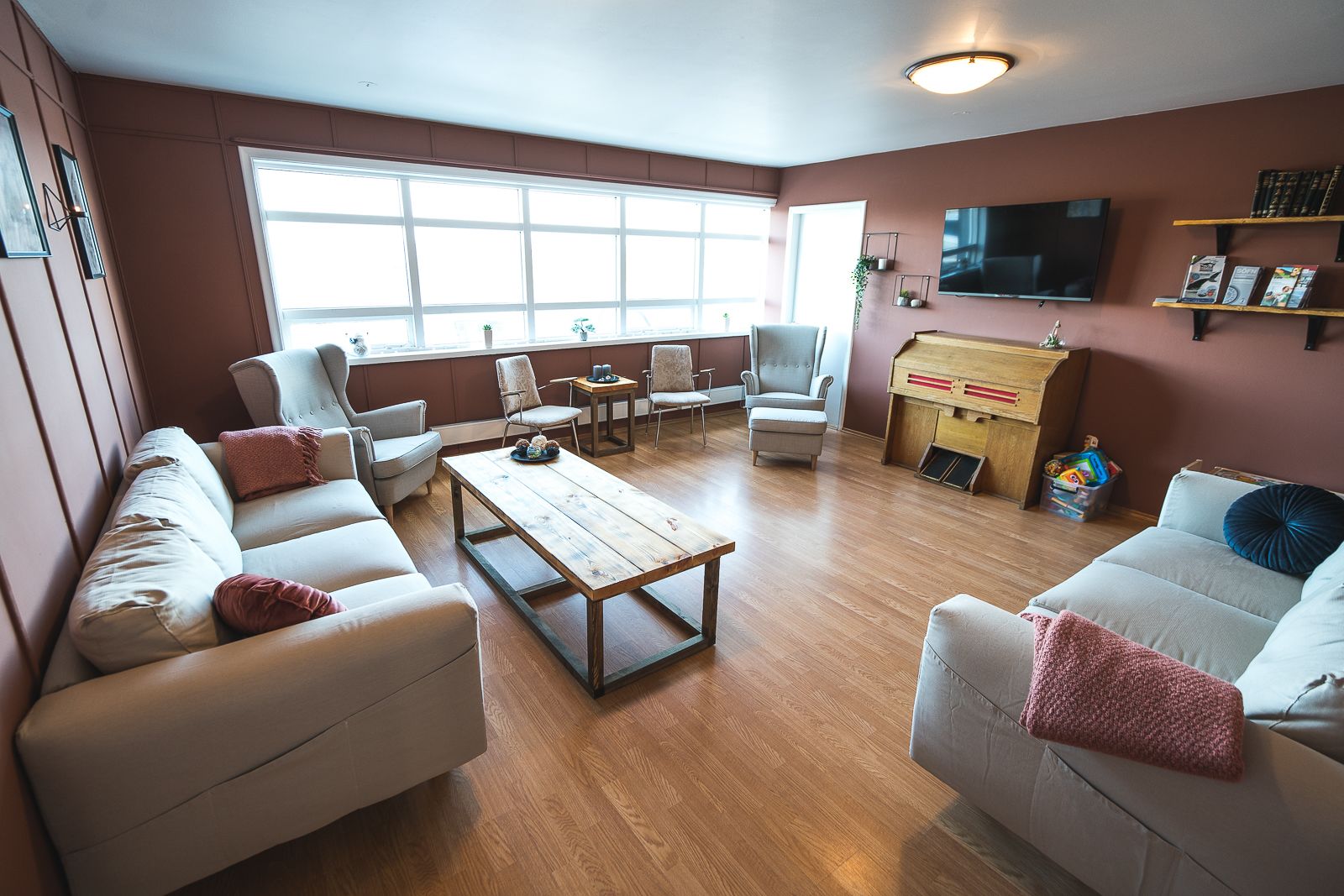 A spacious living room with a wooden coffee table, multiple sofas, and armchairs. There is a television mounted on the wall and a bookshelf with books and decorative items.