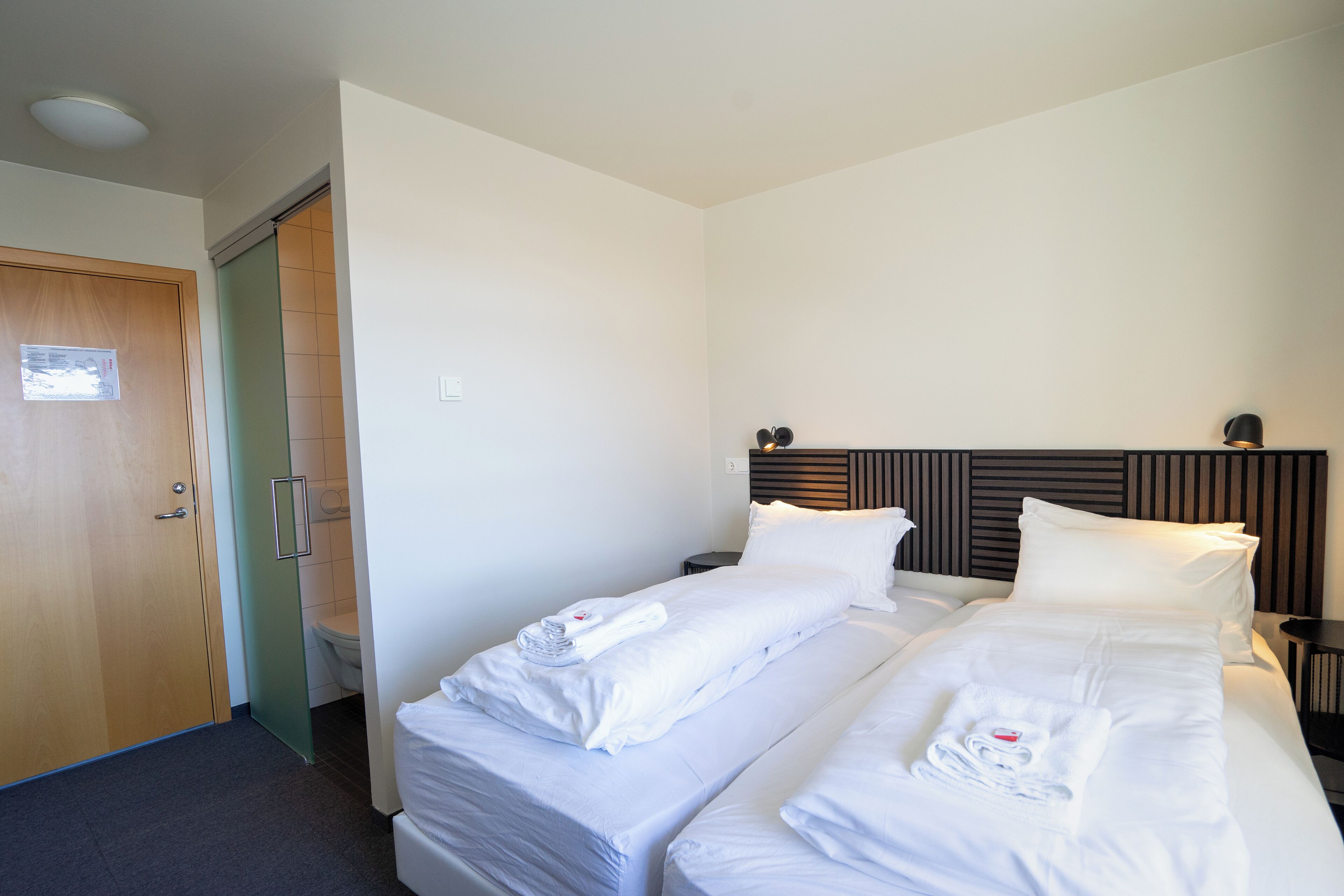 Una camera d'albergo con due letti, una porta in legno, un water e un comodino con una lampada.