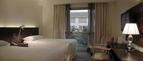 A hotel room with two beds, a desk with a lamp, a chair, and a view of buildings through the window.