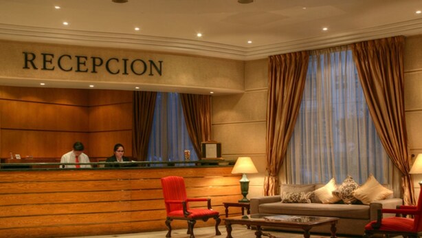 A reception area with a wooden reception desk, two receptionists, a sofa, and a coffee table. - Hotel Costa Real (La Serena)