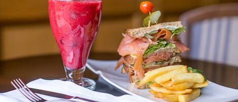 Un repas composé d’un sandwich au saumon fumé, d’une portion de frites et d’un verre d’une boisson rouge, peut-être un smoothie ou un jus de fruits, sur une table avec une serviette blanche et des couverts.