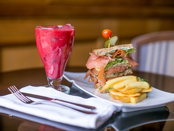Un plato que incluye un sándwich de salmón ahumado, una guarnición de papas fritas y un vaso de una bebida roja, posiblemente un batido o jugo, sobre una mesa con una servilleta blanca y cubiertos.