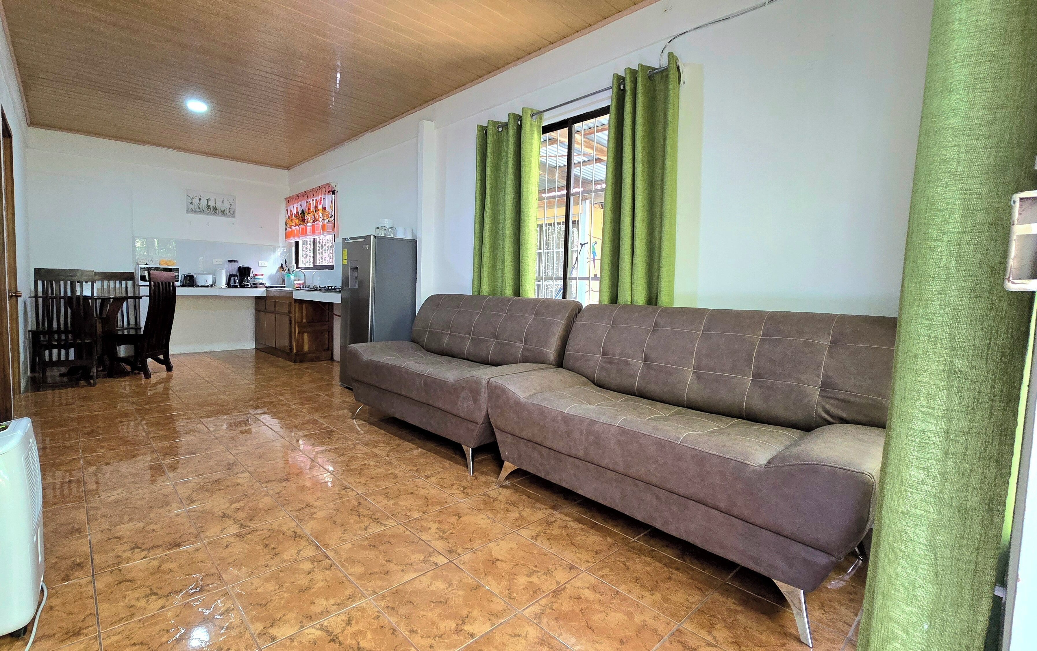 Una sala de estar con sofá, un comedor con sillas, una cocina con electrodomésticos y una ventana con cortinas.