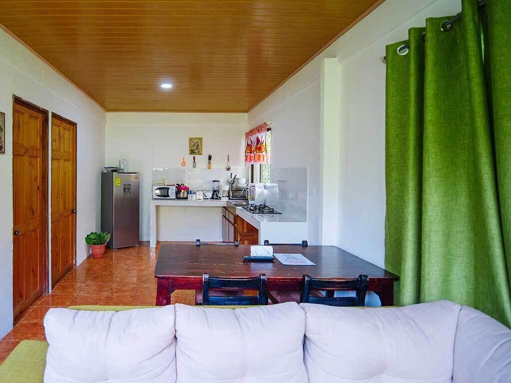 A cozy living room with a wooden ceiling, a dining area with a wooden table and chairs, and a kitchen in the background.