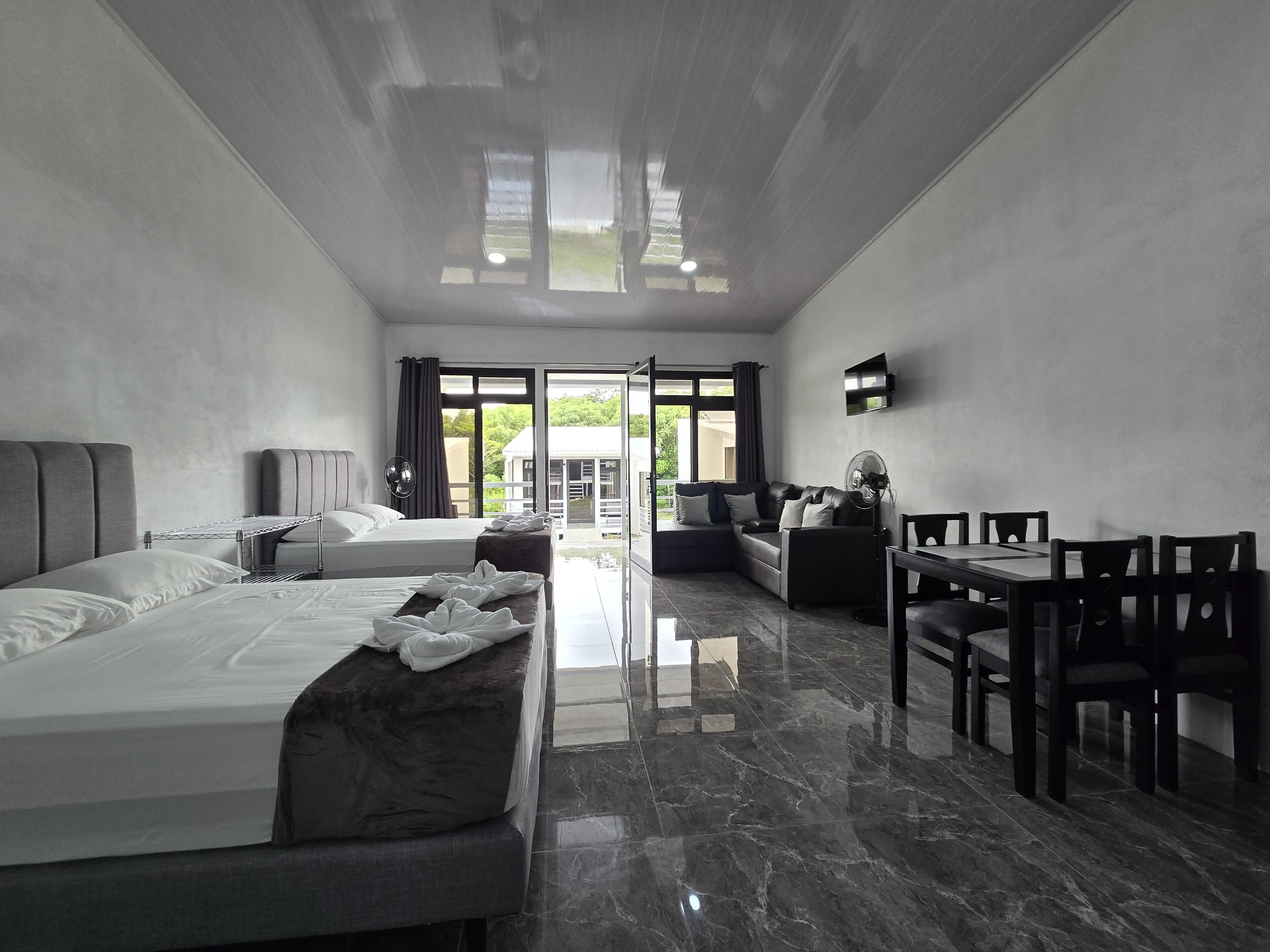 A modern hotel room with a large bed, a sofa, a dining table with chairs, and a TV mounted on the wall.