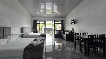 A modern hotel room with a large bed, a sofa, a dining table with chairs, and a TV mounted on the wall.