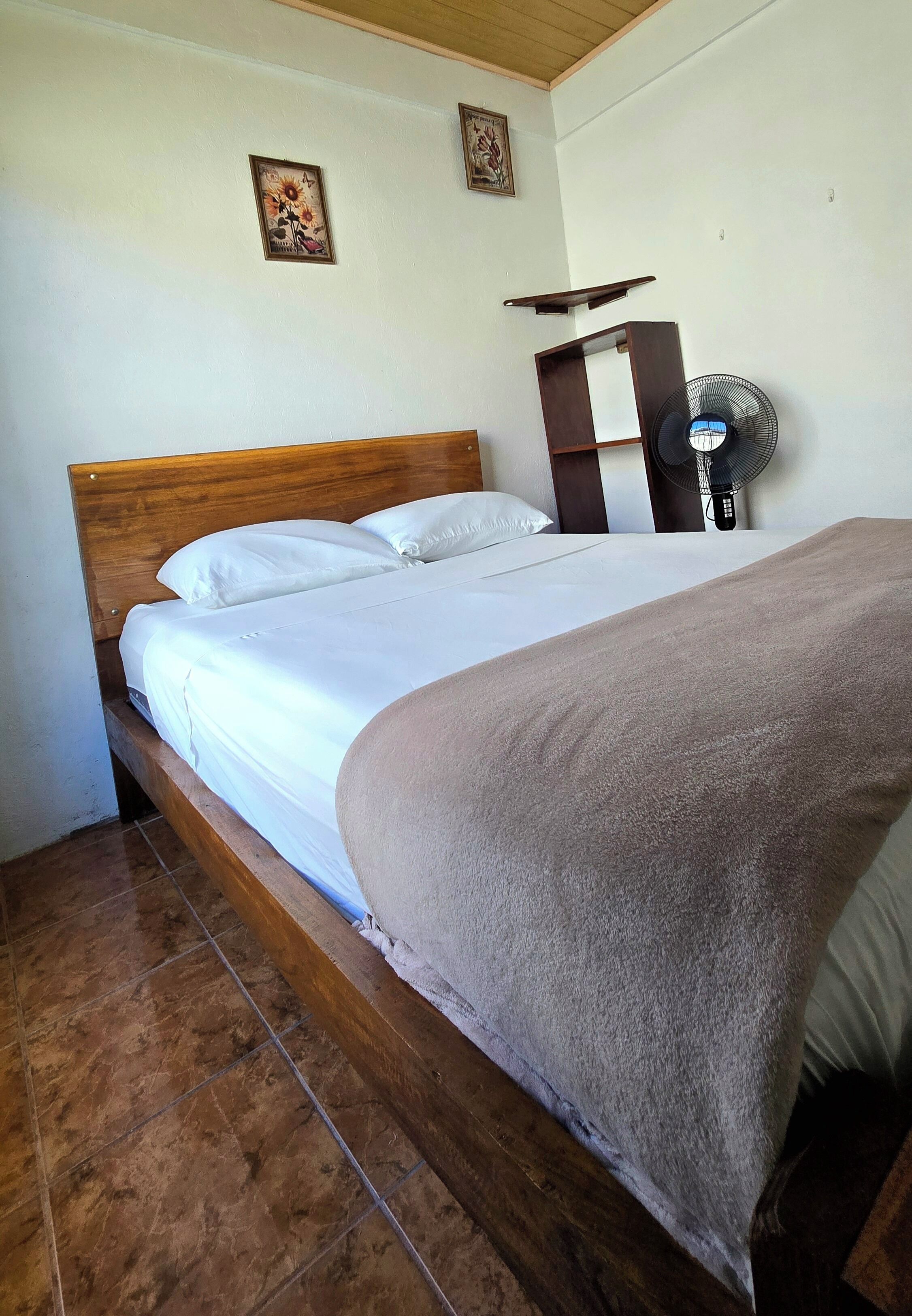A bed with a wooden headboard, white bedding, and a brown blanket. There is a wall-mounted shelf and a standing fan.