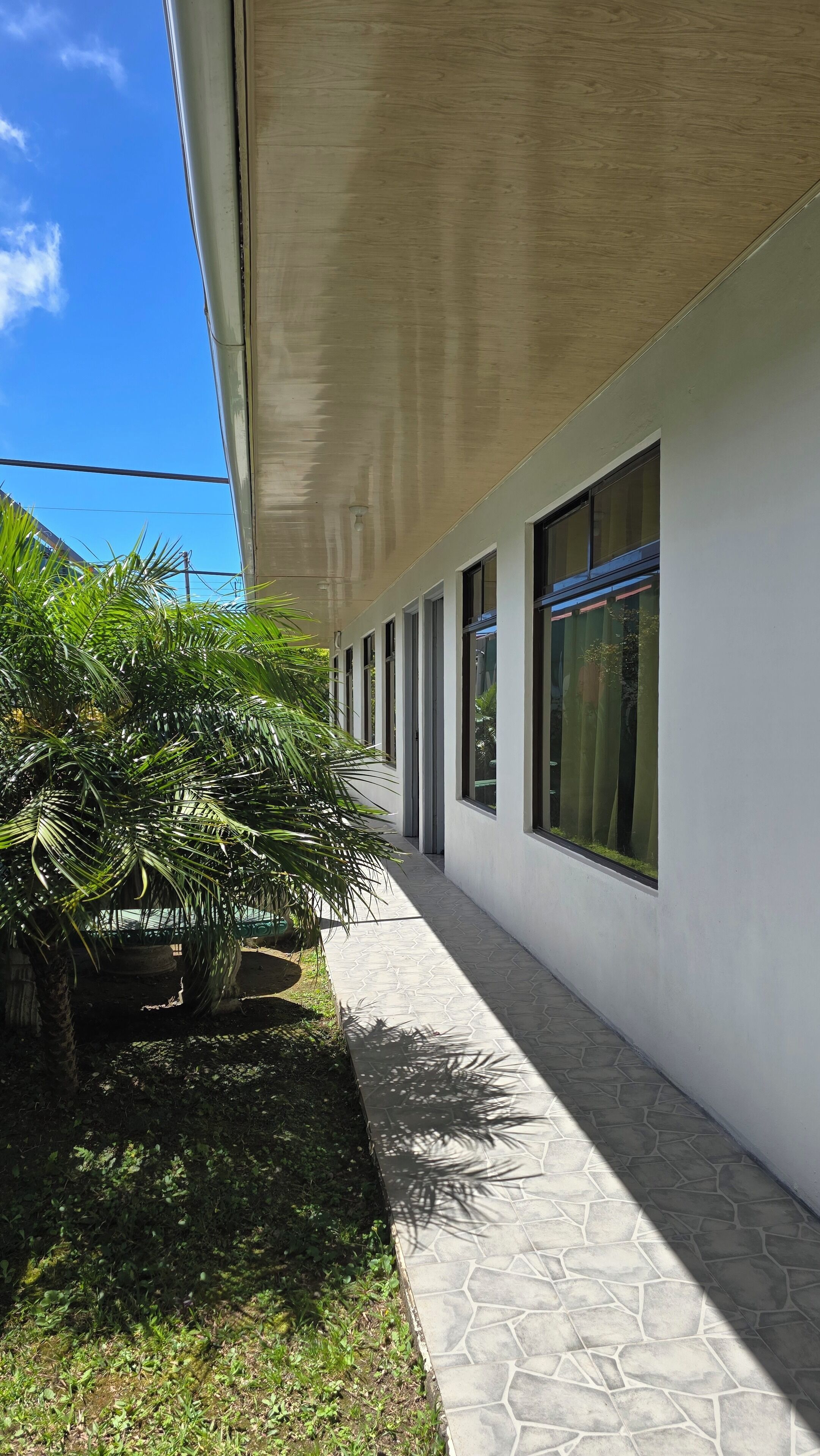 Un pasillo exterior cubierto con suelo de baldosas, ventanas amplias y plantas en macetas.