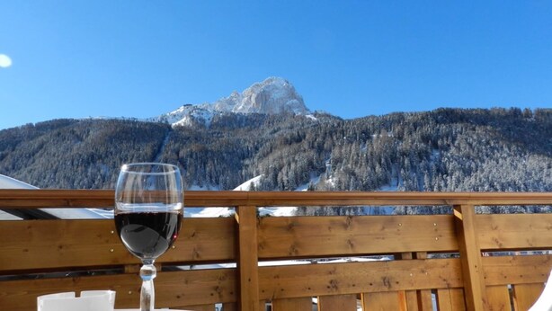 A wooden deck with a glass of red wine, overlooking a mountainous landscape with a clear sky. - Garni Apartments Mezdi (Selva di Val Gardena)