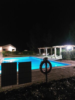 A swimming pool at night with a safety ring, a covered area, and a building in the background.