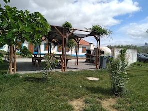 A covered outdoor seating area with a barbecue grill, surrounded by greenery and a building with blue trim in the background.