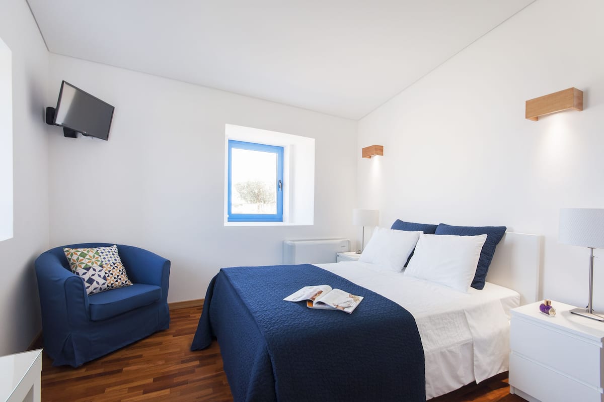 A bedroom with a bed, bedside tables, a chair, a TV mounted on the wall, and a window.