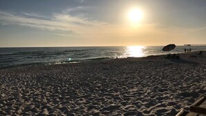 Ein Strand mit einem Pier in der Ferne, bewölkter Himmel und ein Sonnenuntergang über dem Ozean.