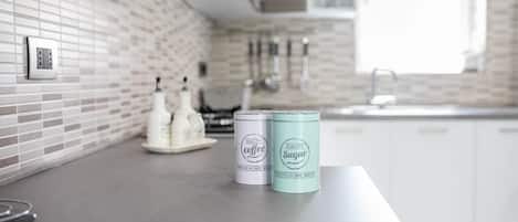A kitchen with a tiled backsplash, a countertop with two containers labeled 'Quality Coffee' and 'Quality Sugar,' and a window providing natural light.