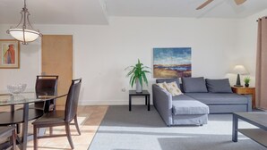 Ein modernes Wohnzimmer mit einem Glastisch, dunklen Holzstühlen, einem grauen Sofa, einem kleinen Beistelltisch mit einer Lampe und einem Deckenventilator.