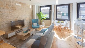 A modern living room with a stone wall, a flat-screen TV, a blue armchair, a sofa, a dining table with chairs, and a large window with a view of buildings.