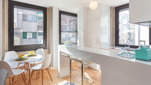 A modern kitchen with a white countertop, bar stools, and a view of the city through large windows.