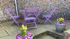Eine Terrasse mit lila Stühlen und Tischen, einer Steinmauer sowie zwei Blumentöpfen mit Narzissen und violetten Blumen.