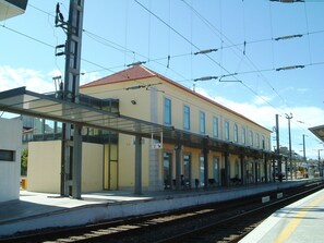 Ein Bahnsteig mit überdachtem Wartebereich, Stromleitungen darüber und einem Gebäude mit roten Ziegeldach.