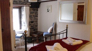 A bedroom with a stone wall, a bed with a red blanket, a chair, a desk, and a mirror.