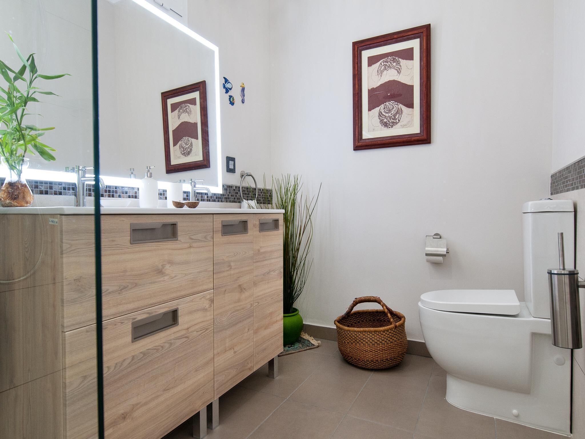 A modern bathroom with a wooden vanity, a sink, a toilet, and two framed artworks on the wall.