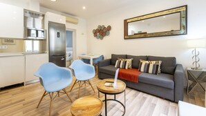 Un espace de vie moderne avec un canapé gris, des chaises bleues, une table basse en bois et un coin cuisine doté d’un réfrigérateur en acier inoxydable.