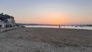 A beach with a row of buildings on the left, a calm sea, and boats in the distance.