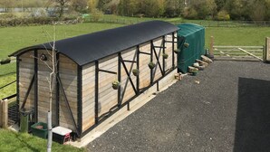 A stable with a black roof and wooden walls, surrounded by a gravel driveway and a grassy field. - Greenacres Self Catering Railway Wagon-Glamping (Bridport)