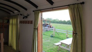 A room with a wooden ceiling, a window with green curtains, a wooden bench, and a view of a tractor in a field. - Greenacres Self Catering Railway Wagon-Glamping (Bridport)