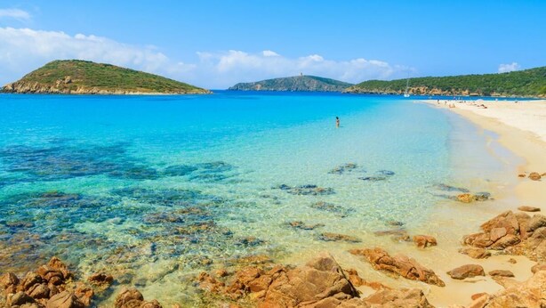 Ein Strand mit klarem türkisblauem Wasser, felsigem Vordergrund und einem sandigen Strandabschnitt mit Menschen.