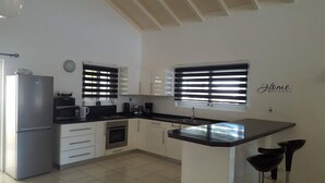 Una cocina moderna equipada con refrigerador, estufa, microondas y cafetera. Hay dos ventanas con persianas, un fregadero y una encimera con sillas.