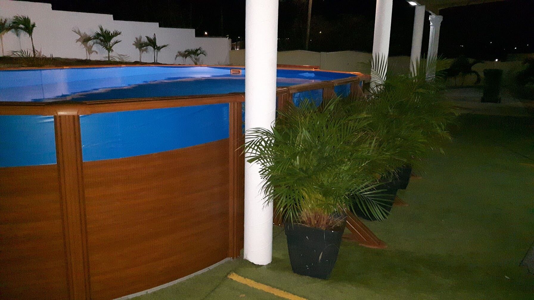 A poolside area with a wooden pool deck, a potted plant, and a white column.