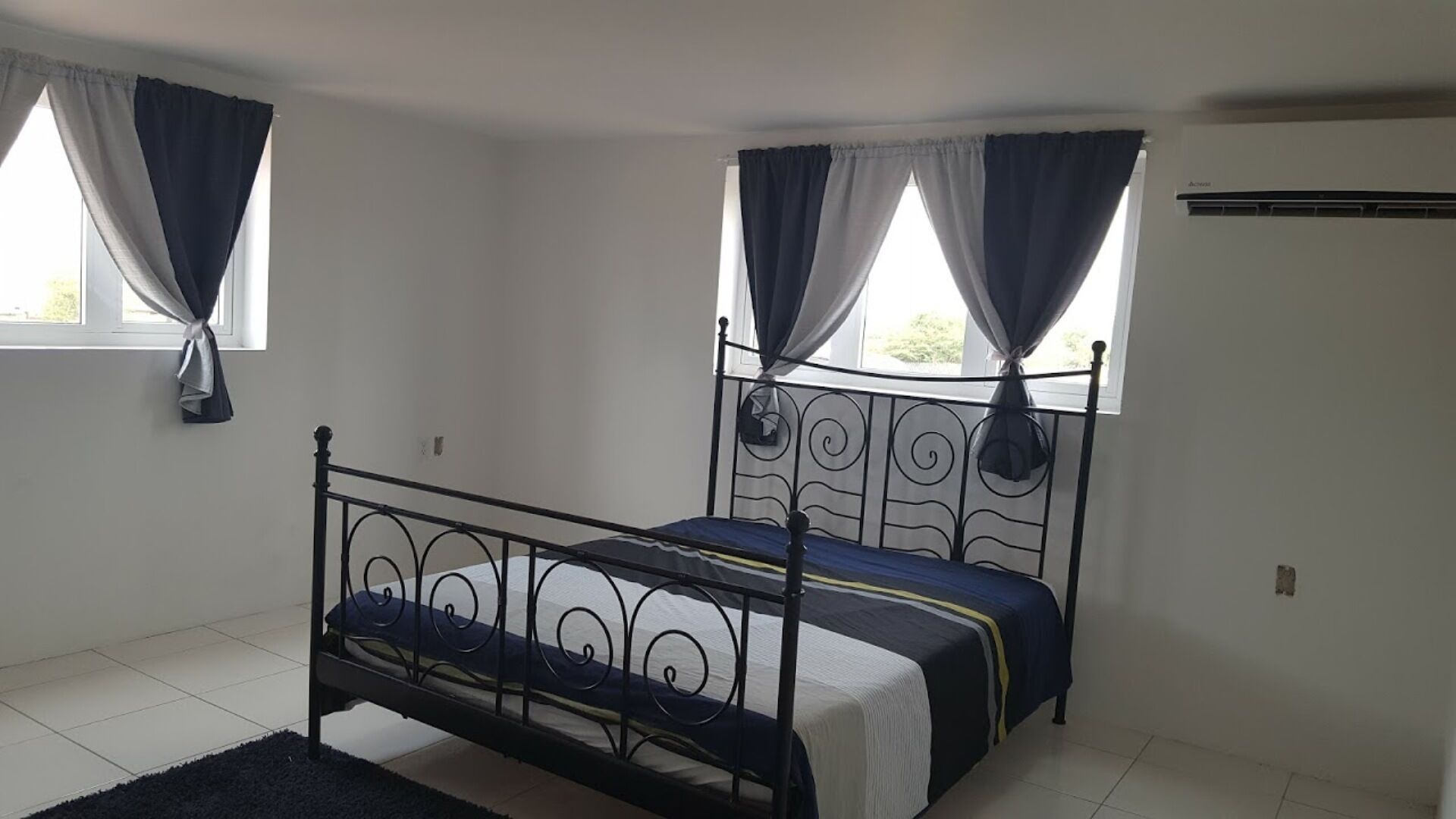 Una cama individual con estructura metálica y colchón oscuro en una habitación con paredes blancas y piso de baldosas.