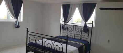 Una cama individual con estructura metálica y colchón oscuro en una habitación con paredes blancas y piso de baldosas.