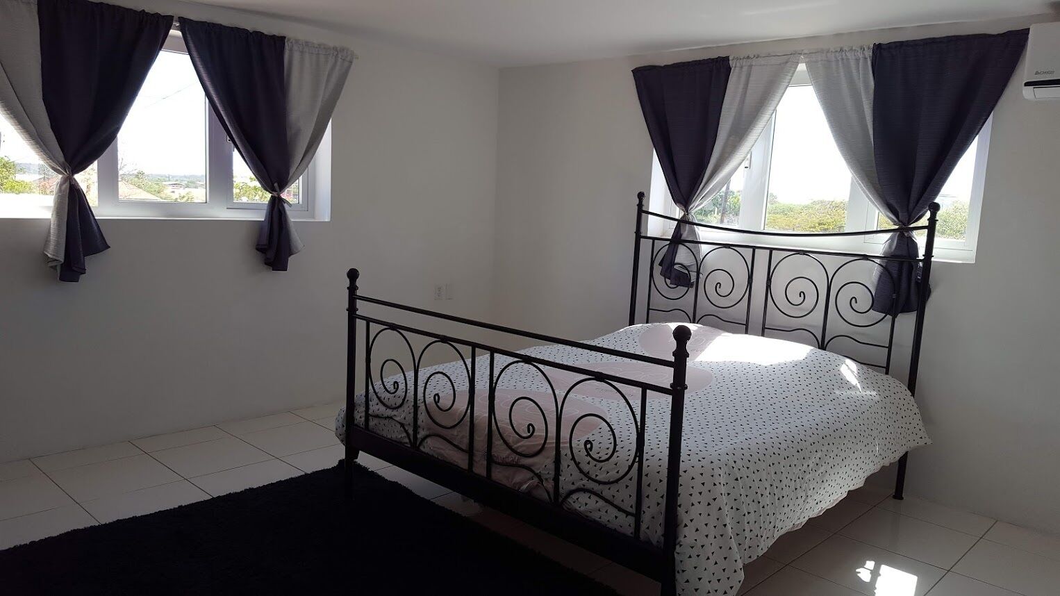 A single bed with a metal frame and a white bedspread with black dots, positioned in a room with white walls and a black rug.