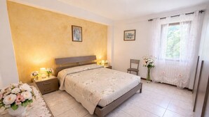 A bedroom with a bed, bedside tables, a chair, a window with curtains, and two wall-mounted pictures. - Casa Rosalia patio apartment (Tremosine)