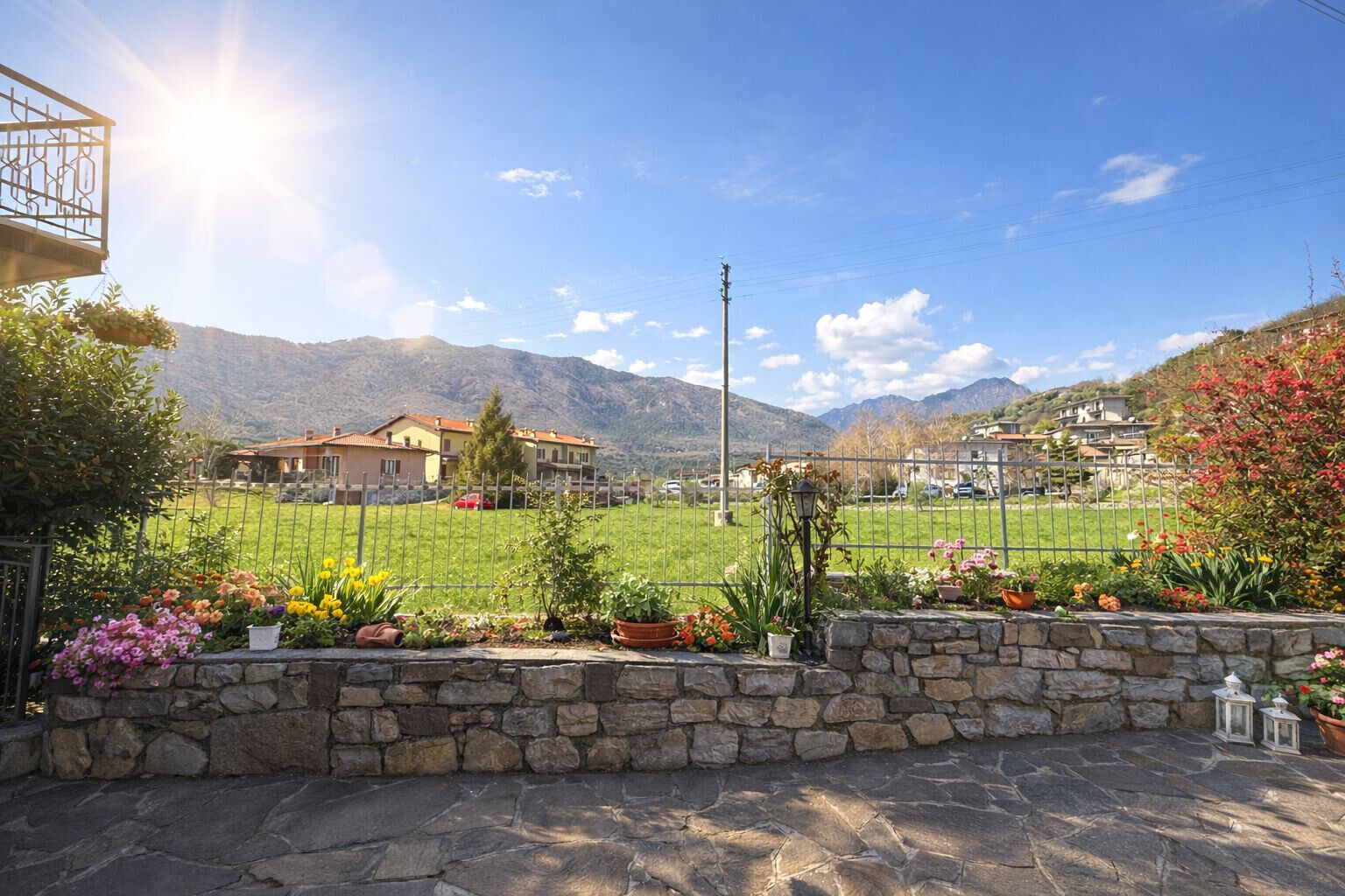 A stone wall with potted plants, a metal fence, and houses in the background.