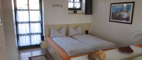 A bedroom with a bed, a bedside table, a window with curtains, a framed picture on the wall, and a door.