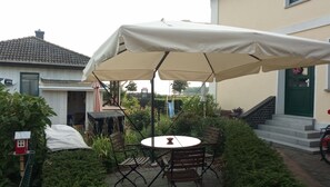 Un patio con tavolo e sedie, riparato da un ampio ombrellone, circondato da piante e da una casa.