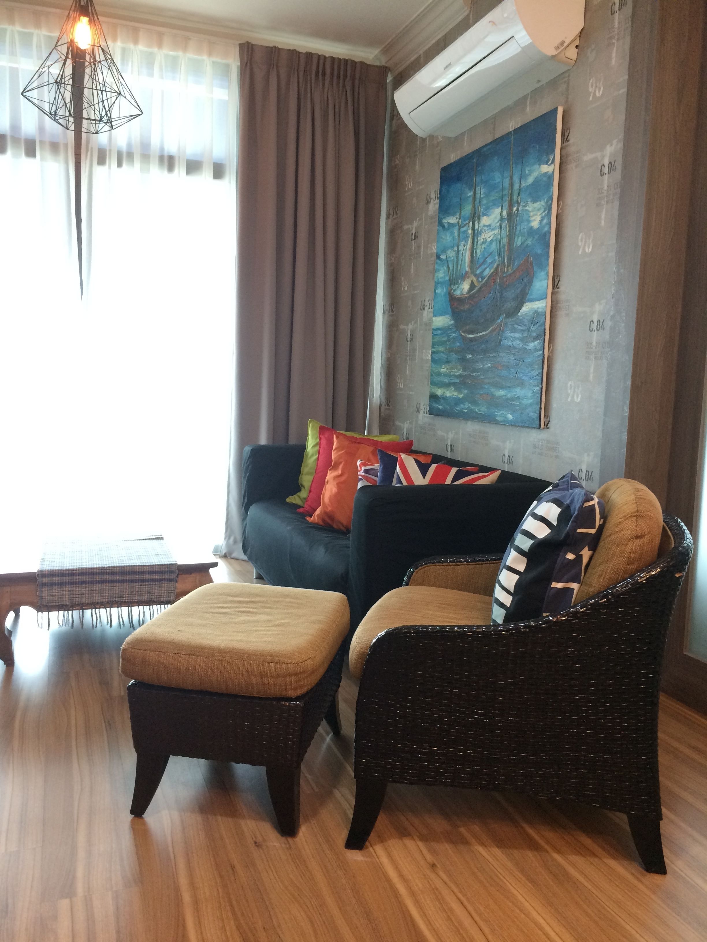 A living room with a sofa, ottoman, and wicker chair, a painting on the wall, and a hanging light fixture.
