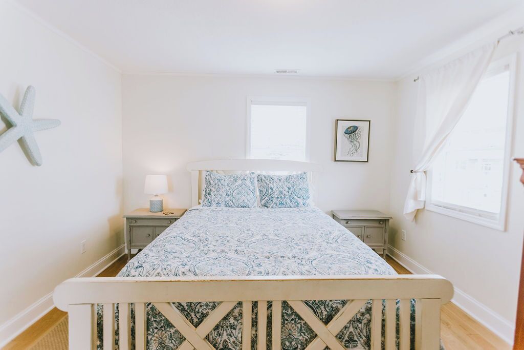 Quarto com cama, mesas de cabeceira, decoração de estrela-do-mar, quadro emoldurado e janela com cortinas.